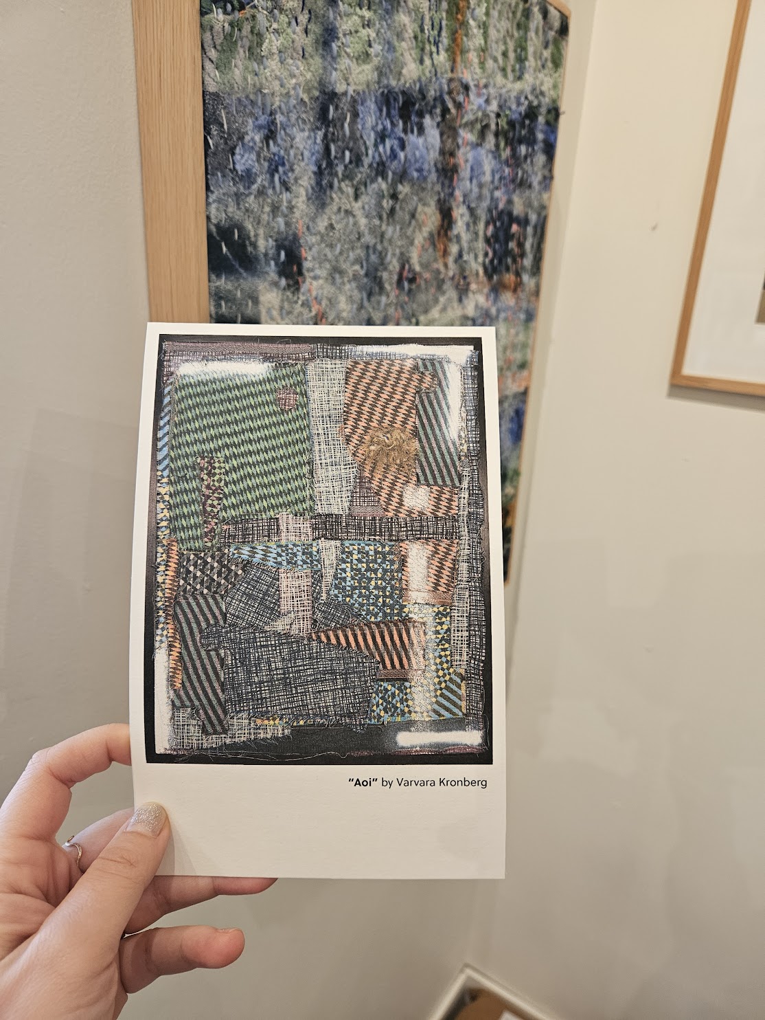 A hand holding the 'Aoi' postcard at an exhibition, a framed textile work on the gallery wall behind