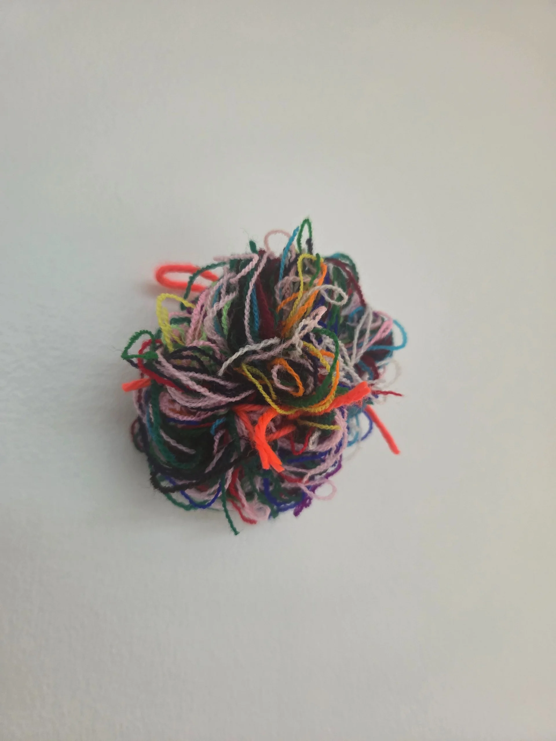 A round brooch made of many tangled leftover yarns in every colour, on a white wall
