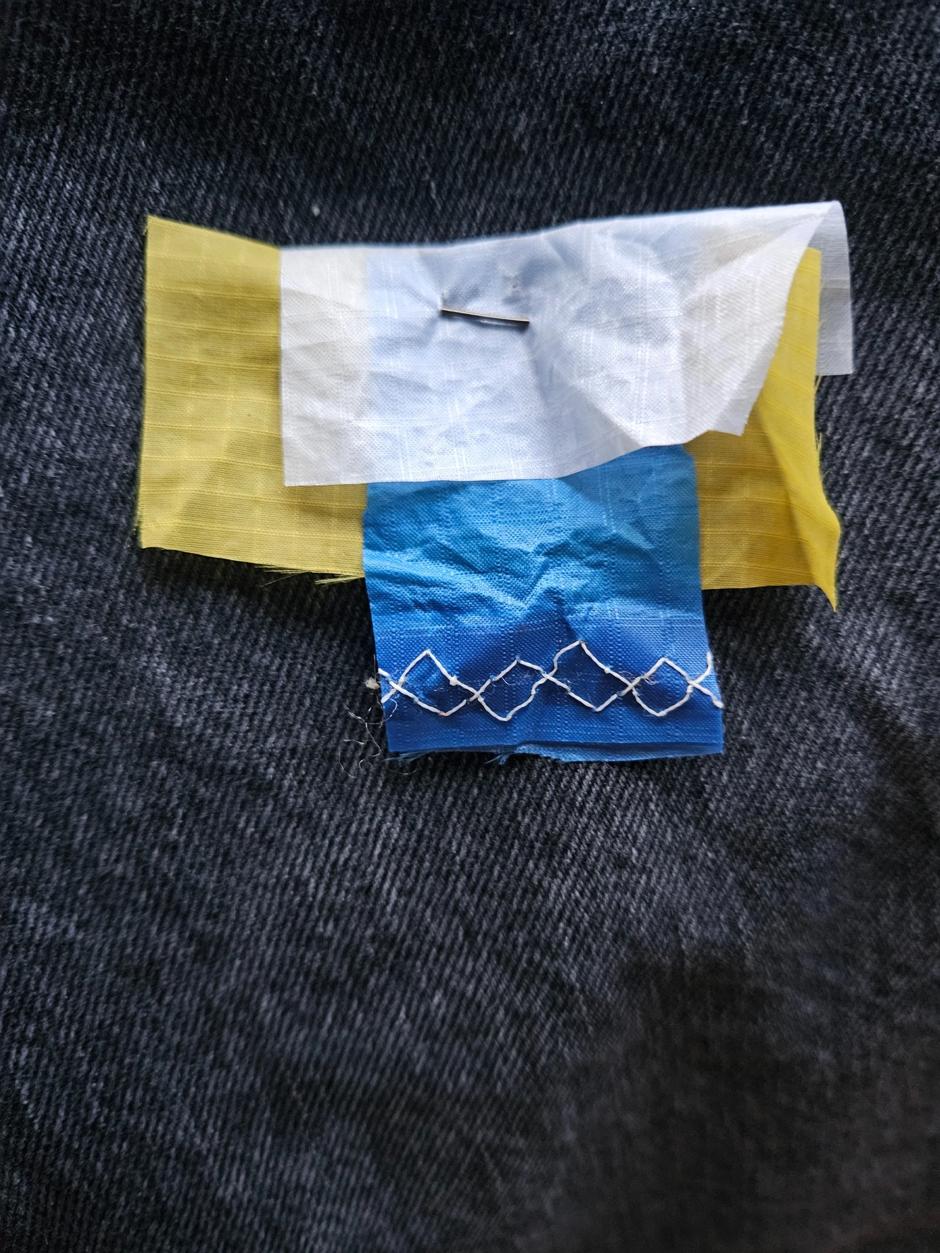An "I care" pin — a small patch of blue, yellow and white reclaimed sailcloth with white zigzag stitching, on denim