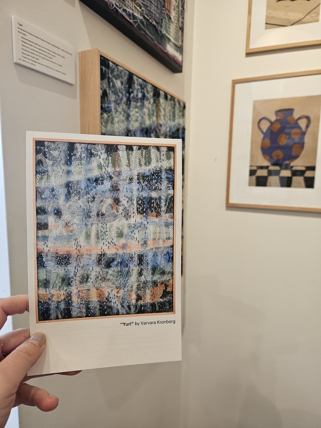 The framed work on a gallery wall, with the artwork's printed postcard held in front of it